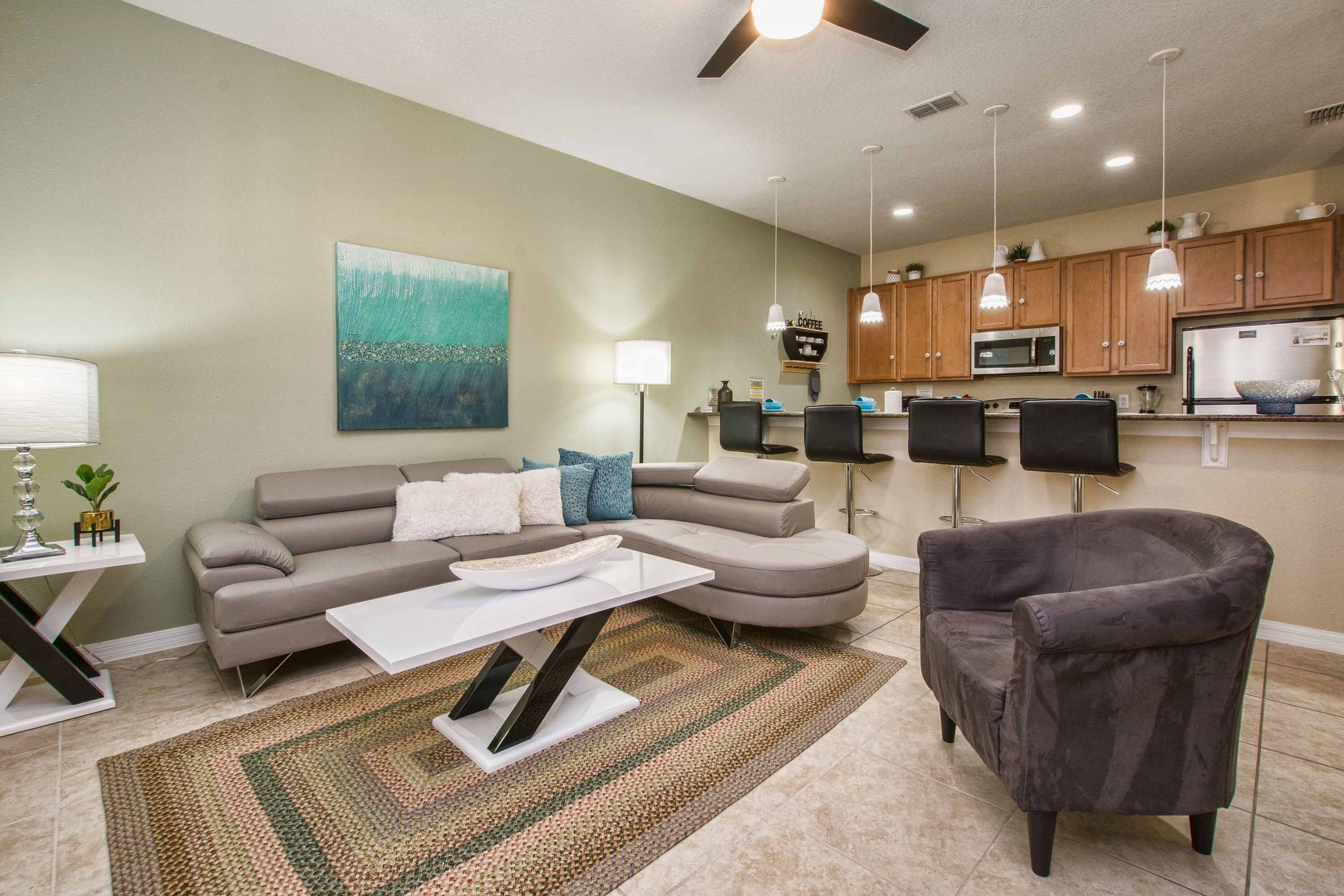 A living room with a sofa, coffee table, and armchair, a kitchen with wooden cabinets and appliances, and a ceiling fan.