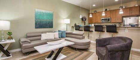 A living room with a sofa, coffee table, and armchair, a kitchen with wooden cabinets and appliances, and a ceiling fan.