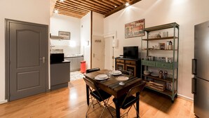 A compact kitchen with a dining area, wooden ceiling beams, a TV, and a green metal shelving unit with kitchenware.