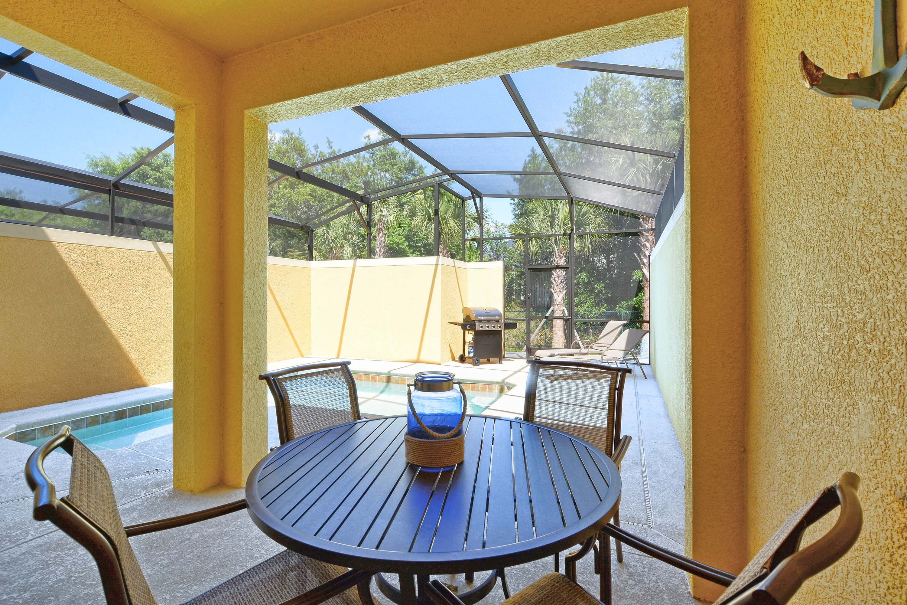 A covered outdoor patio with a round table and chairs, a grill, and a pool in the background.