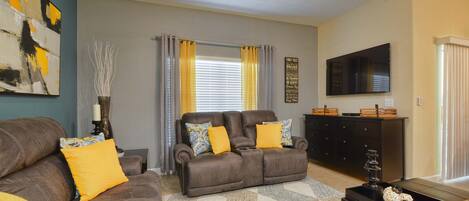 A living room with a brown sofa set, a television mounted on the wall, and a ceiling fan.