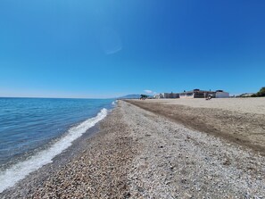 Une plage de galets, un ciel dégagé, quelques bâtiments visibles au loin et une mer calme.