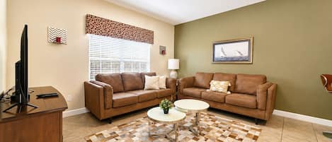 A living room with two brown leather sofas, a coffee table, a TV on a wooden stand, a window with blinds, and a framed picture on the wall.