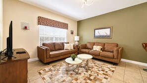 A living room with two brown leather sofas, a coffee table, a TV on a wooden stand, a window with blinds, and a framed picture on the wall.