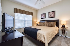 Quarto de hotel com uma cama, uma televisão sobre uma escrivaninha, duas luminárias fixadas na parede e um ventilador de teto.