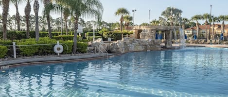 A clear swimming pool with a rock feature, surrounded by palm trees and a building in the background.