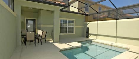A swimming pool with a covered patio area, a table with chairs, and a door leading to an interior space.