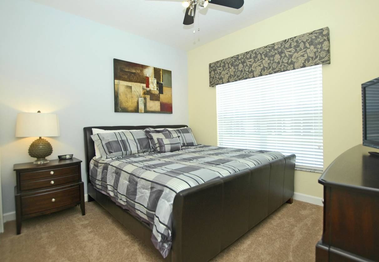 Une chambre à coucher comprenant un lit, une table de chevet, une lampe, un ventilateur de plafond, des œuvres d’art, des stores et une commode.