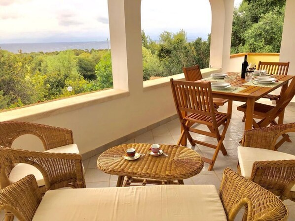 A covered outdoor dining area with wicker furniture and a table set for two, overlooking a scenic landscape.