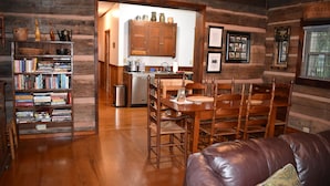 A log cabin interior with a wooden dining table, chairs, a leather couch, a bookshelf, and a kitchen area. - 1850s log cabin 1 mile to downtown Weaverville 15% discount 7 days or more (Weaverville)