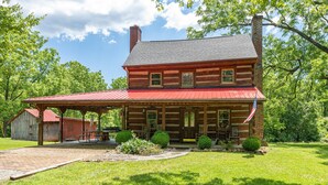 A two-story log cabin with a red metal roof, a porch with benches, and a flagpole with an American flag.