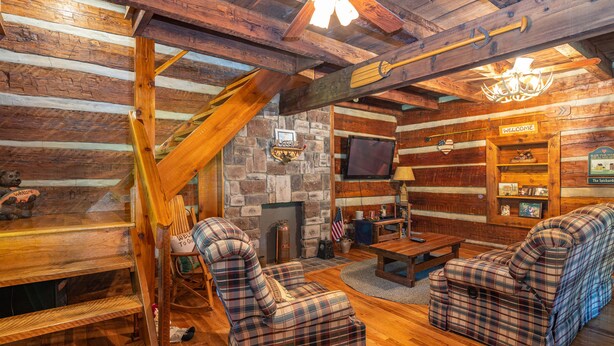 A log cabin interior with a stone fireplace, plaid furniture, and a wooden ceiling.