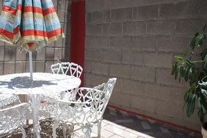 Juego de mesa y sillas de metal blanco frente a un muro de ladrillo, con un paraguas a rayas y una planta en maceta cerca.