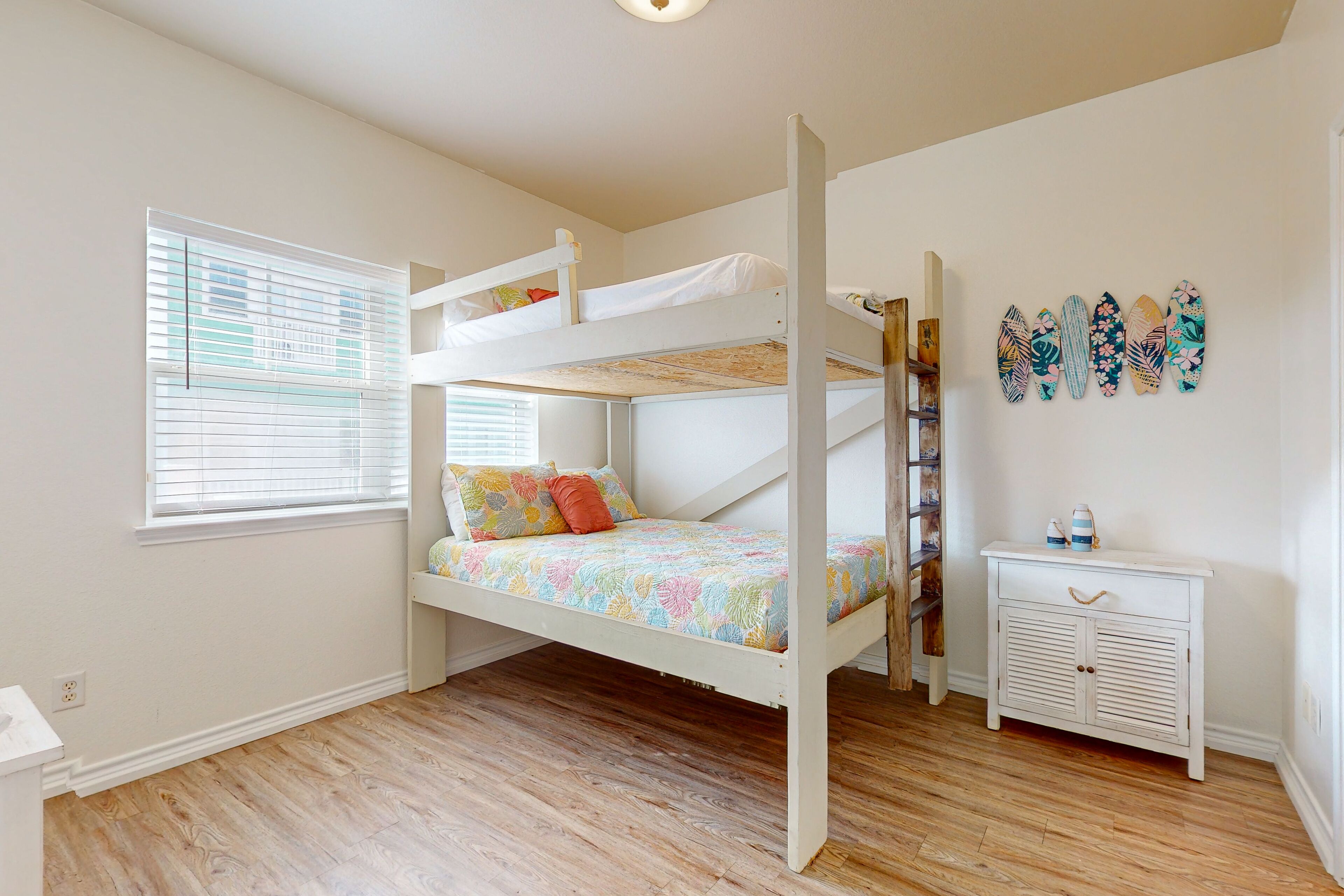 A bunk bed room with a window, a small white dresser, and a wall with decorative items.