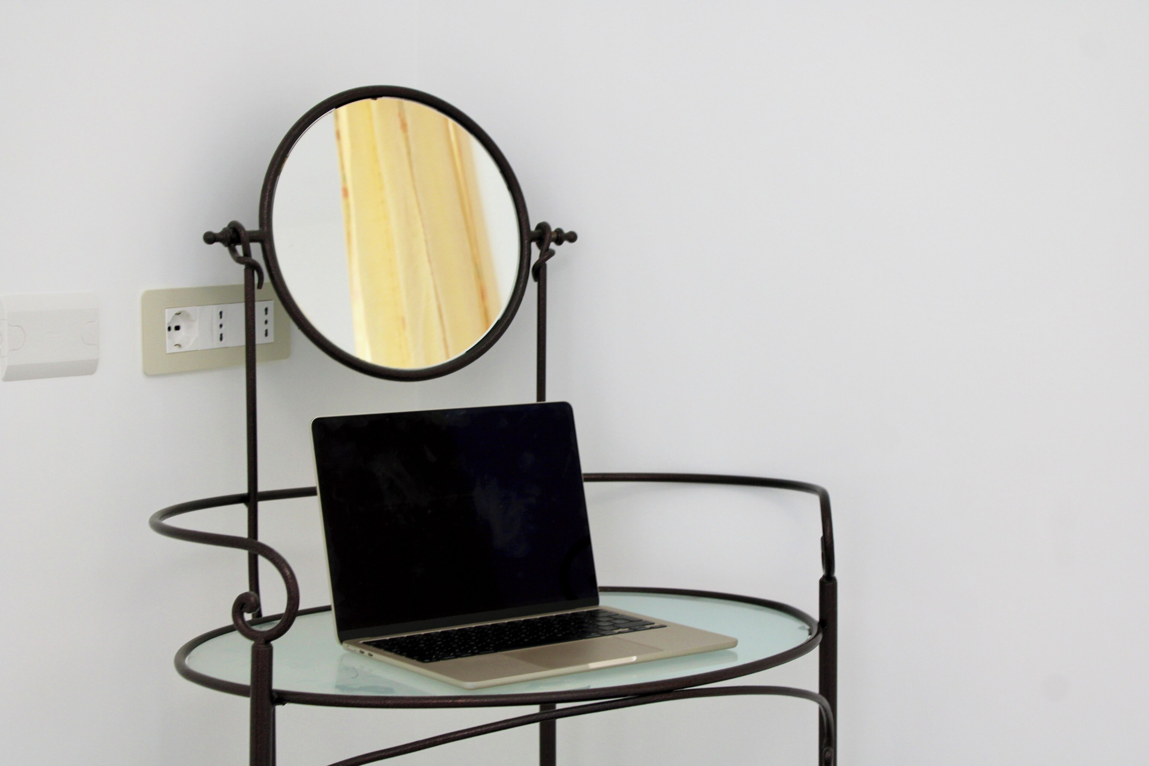 A laptop on a glass table with a black metal frame and a round mirror above it.
