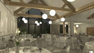 A spacious dining area with high ceilings and large windows, featuring round tables set with white tablecloths and clear glassware.