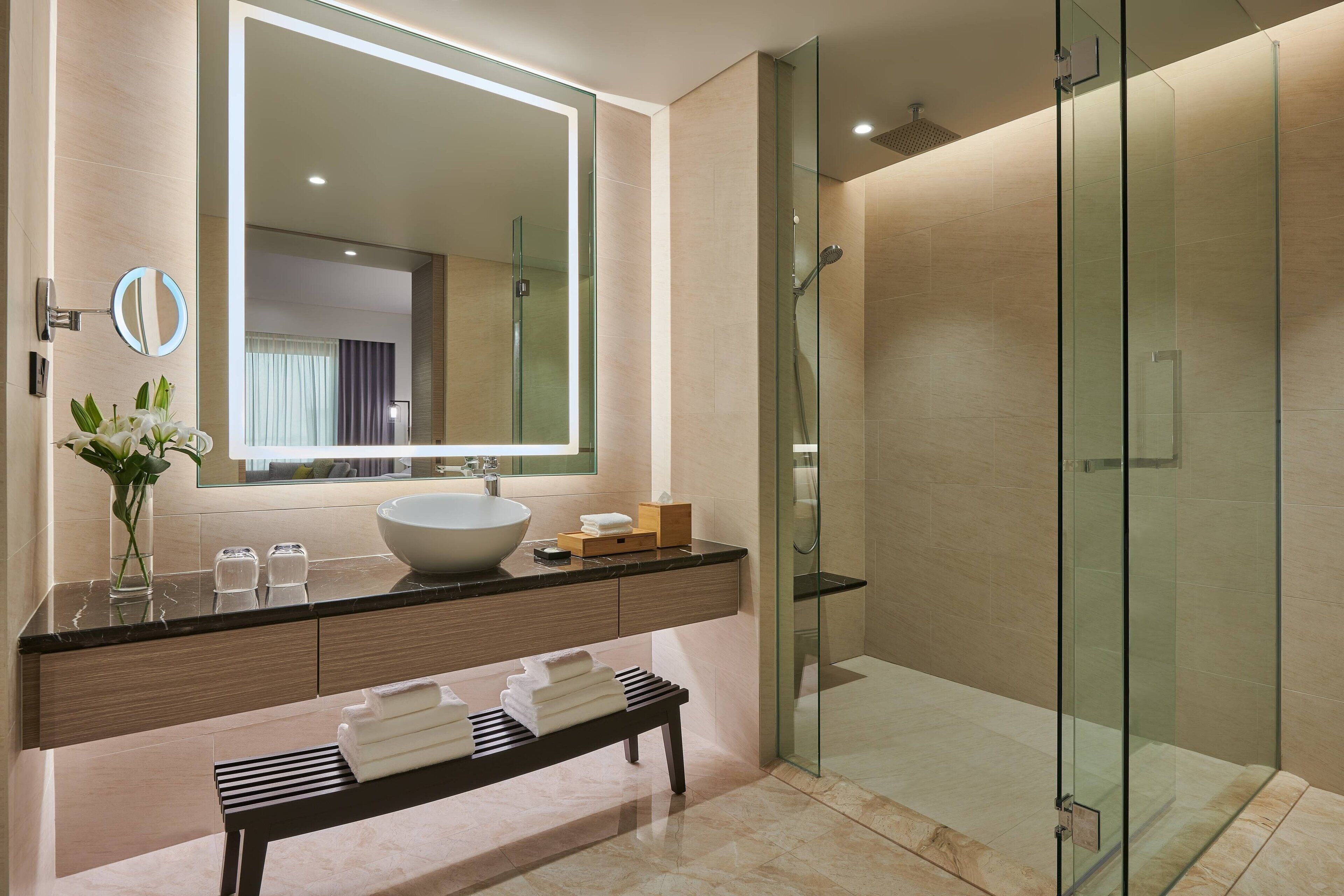 A modern bathroom with a large mirror, a vessel sink, a glass-enclosed shower, and a bench with towels.
