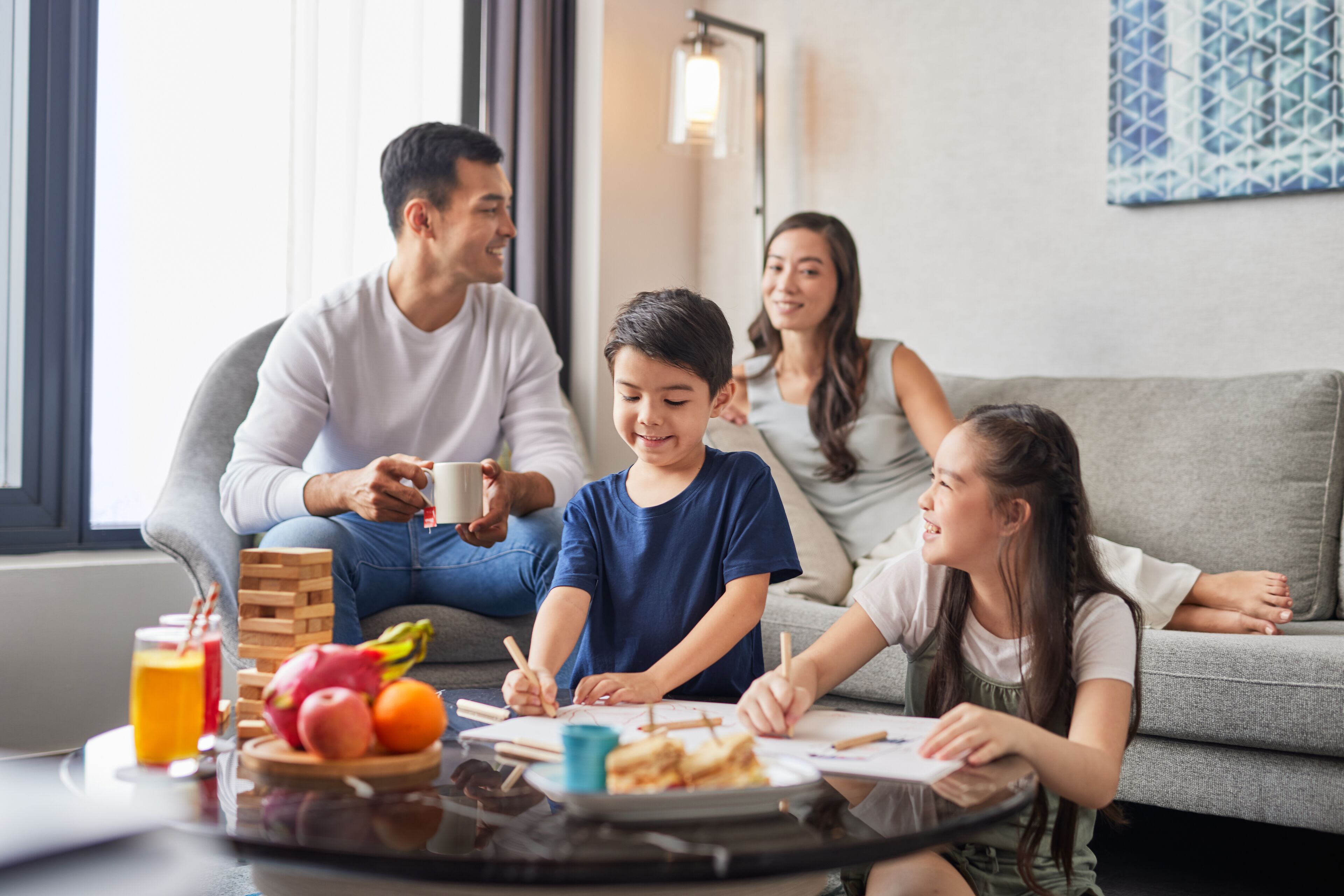 Una familia de cuatro personas sentada en un sofá gris en una sala de estar, con una mesa de centro frente a ellos.