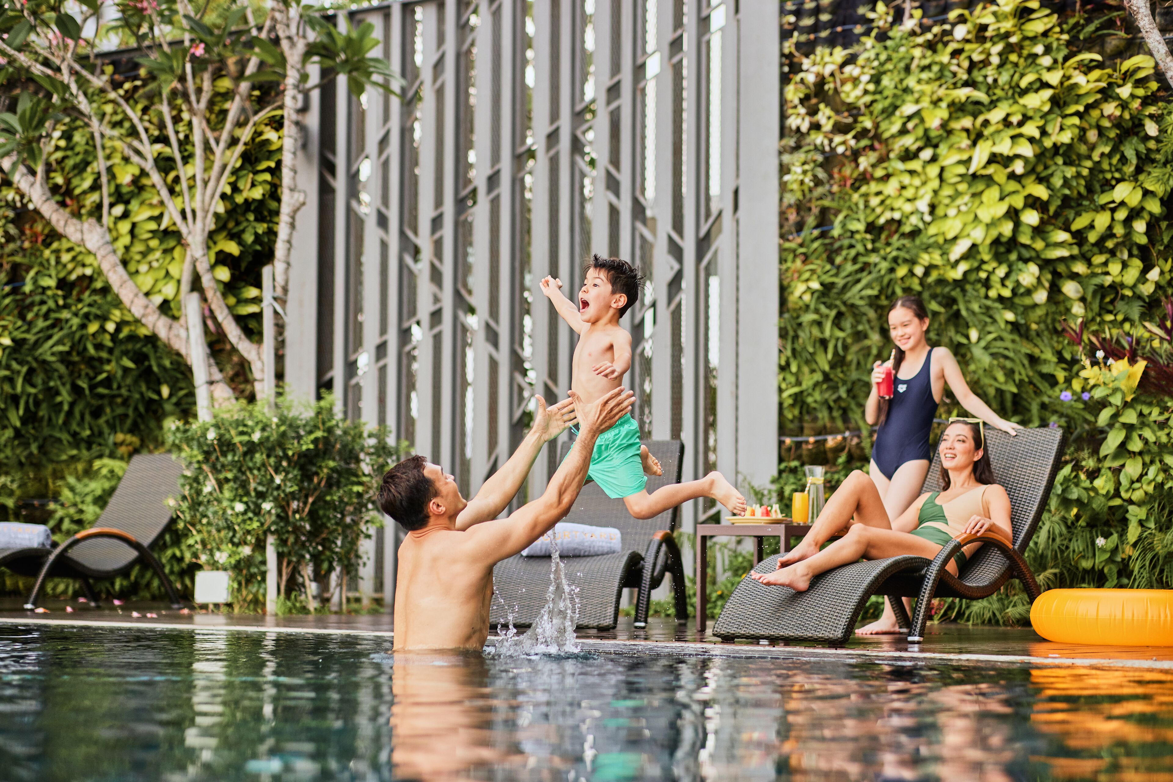 A poolside scene with a man lifting a child, a woman sitting on a lounge chair, and another woman standing by the pool.