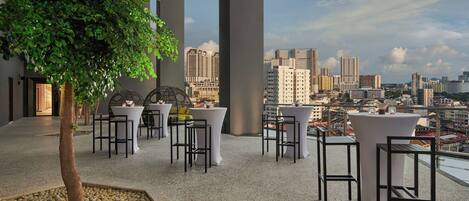 A terrace with tables and chairs, a tree, and a city view.