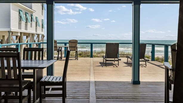 Een houten terras met buitenmeubilair, uitzicht op de oceaan en een gebouw met een balkon.