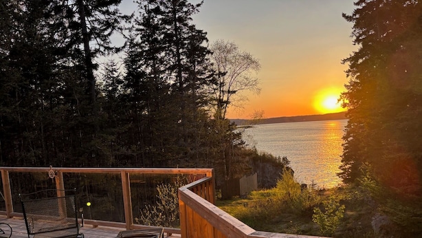 A wooden deck with two metal chairs and a table, overlooking a body of water with a sunset in the background. - Oceanfront cottage with new Oceanfront Master BR in 2023, complete remodel 2021 (Wilsons Beach)