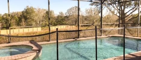 A clear outdoor swimming pool with a brick deck, surrounded by a metal fence and trees in the background.