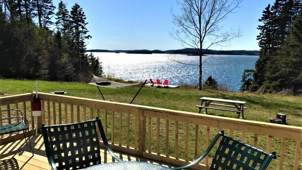 A deck with a glass table and chairs, overlooking a lake with a picnic area in the background. - Starfish Cottage:  Ocean Views, Walking Trail Above the Shore with NEW Master BR (Wilson Beach)