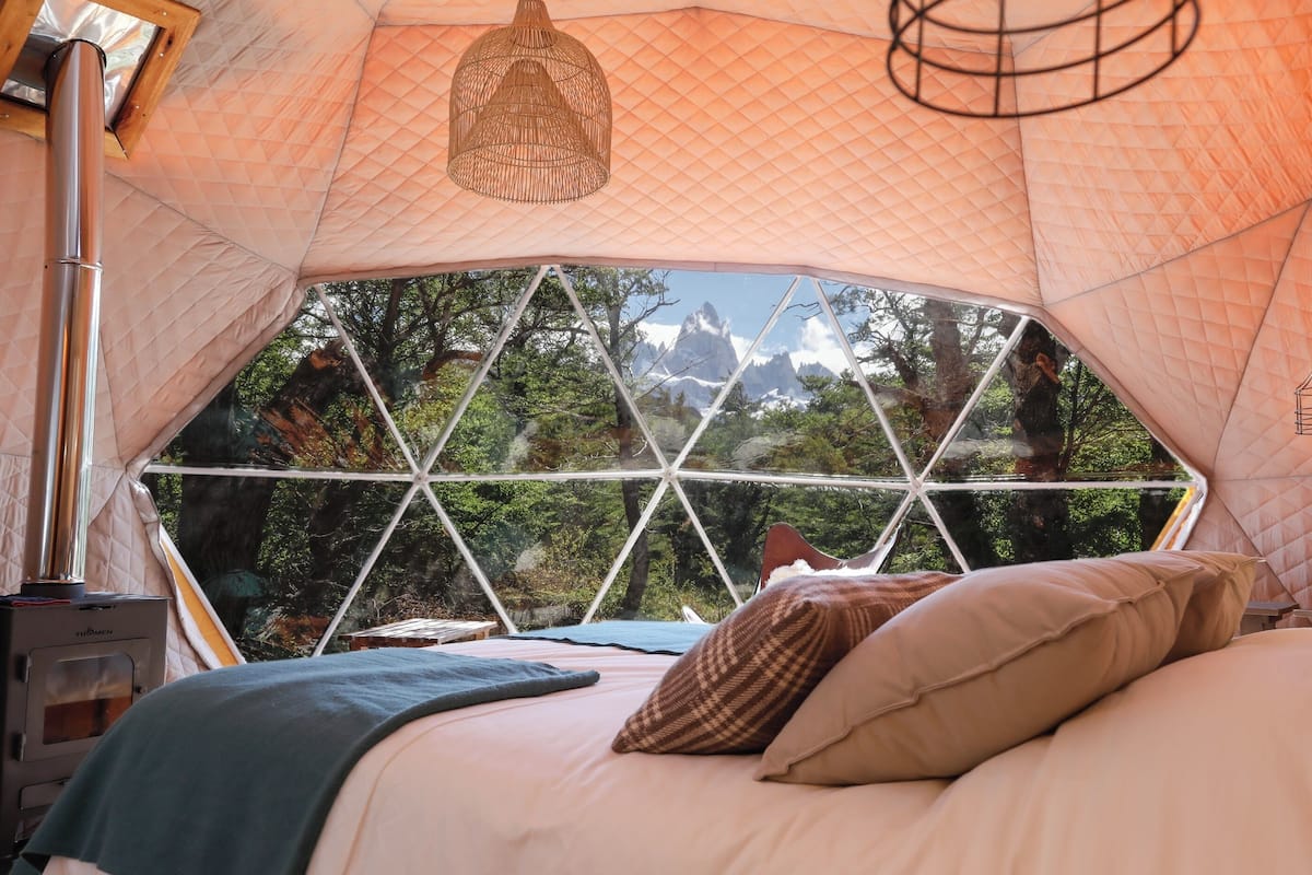 Een knus, achthoekig tentvormig vertrek met een bed, kussens, een lamp en uitzicht op een bergachtig landschap door een geometrisch raam.