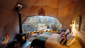 A cozy, well-lit tent with a large window, a bed with pillows, a wooden table, and a fireplace. - Chalten Camp - Glamping With A View (El Chalten)