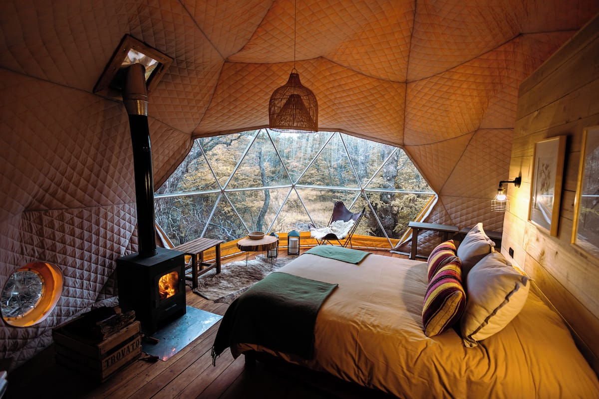 Een gezellige, goed verlichte tent met een groot raam, een bed met kussens, een houten tafel en een open haard.