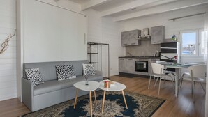 Una sala moderna con un sofá gris, dos mesas redondas blancas y un tapete estampado. Se ve, al fondo, una cocina con armarios grises y una zona de comedor con una mesa y sillas blancas.