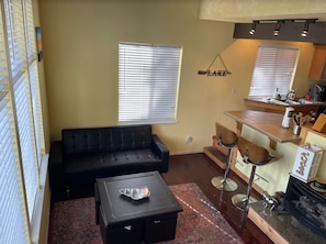 Un espace de vie compact comprenant un canapé en cuir noir, une petite table basse, un coin cuisine avec un bar, et une cheminée.