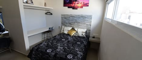 A bedroom with a bed, a desk, a chair, a shelf, and a window.