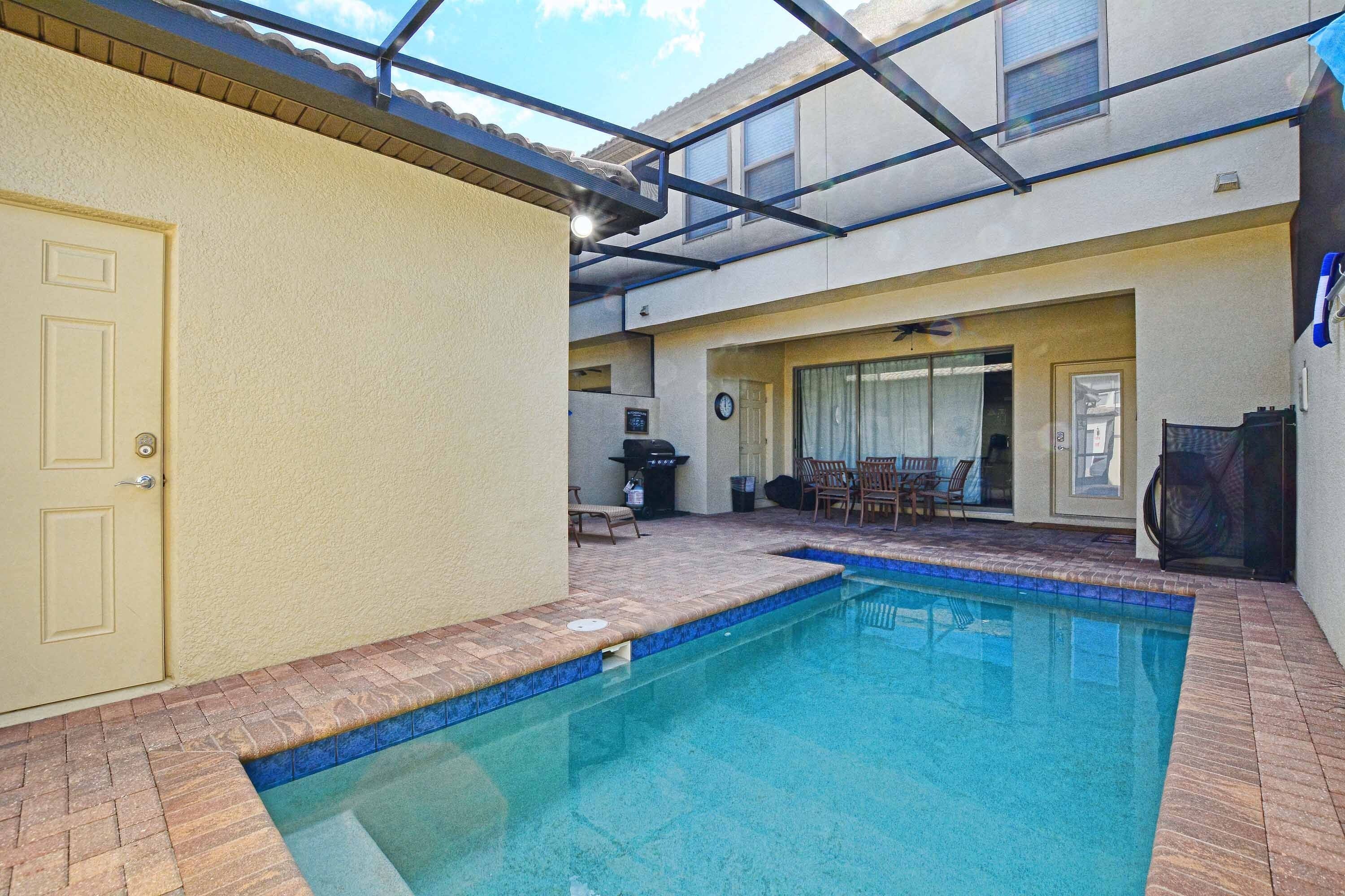 A swimming pool with a tiled edge, a door, and a covered patio area with chairs and a grill.