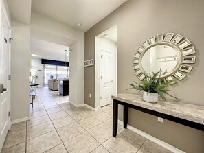 A hallway with a tiled floor, a round mirror with a sunburst frame, a potted plant, and a door with a 'RELAX' sign.