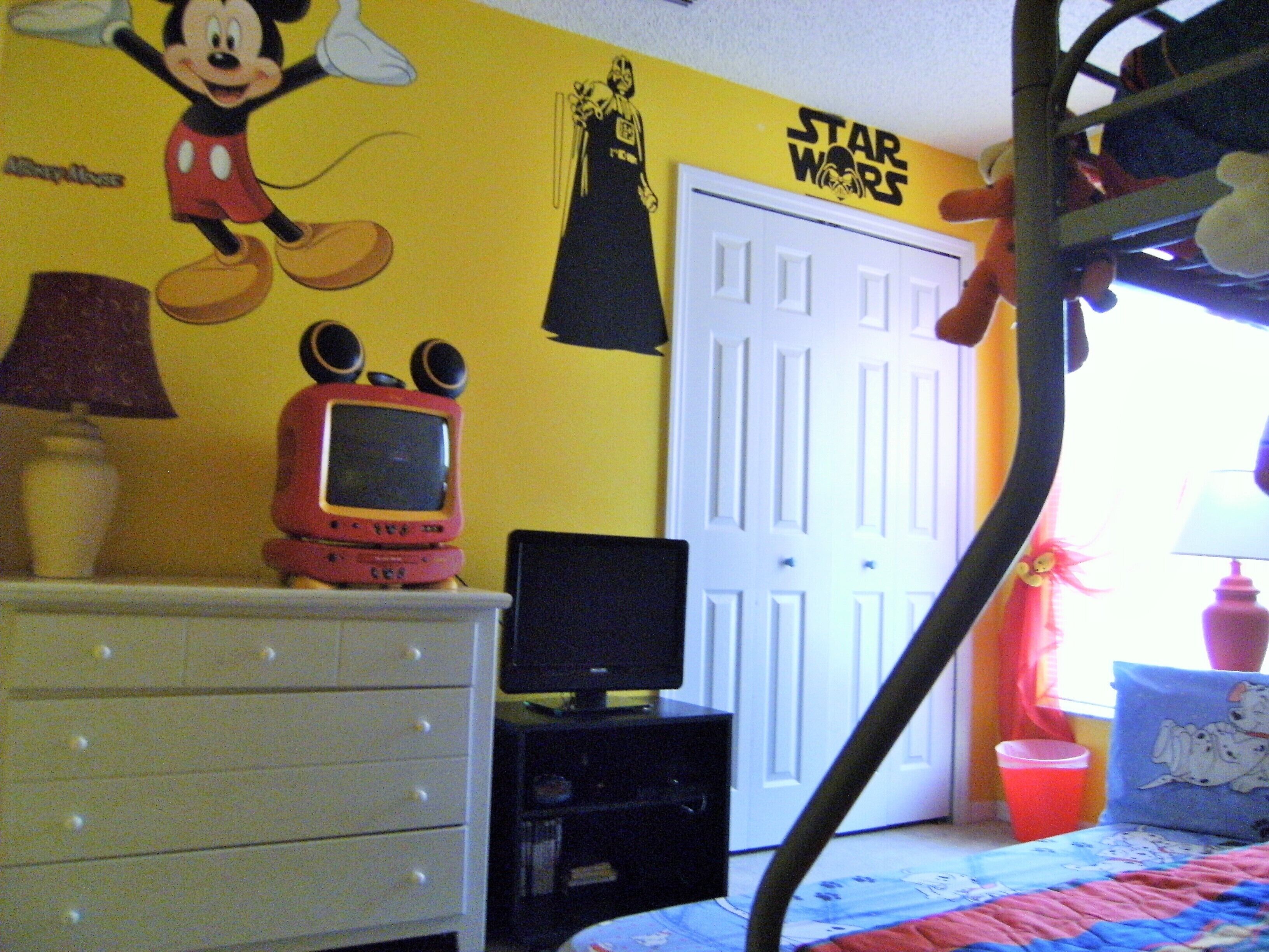 A child's bedroom with a bed, dresser, and wall decorations including a Mickey Mouse and Star Wars theme.