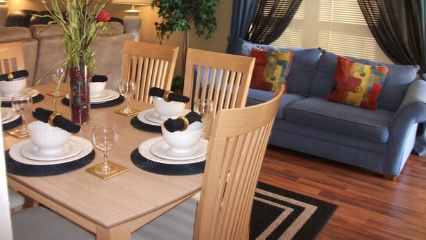 A dining area with a table set for four, surrounded by wooden chairs. There is a vase with flowers on the table, and a plant is visible in the background. - Prime Location In Secure Safe 247 Manned Gatehouse Community With Clubhouse (Kissimmee)