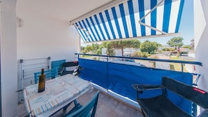 Ein Balkon mit einer blau-weiß gestreiften Markise, einem Tisch mit einer Flasche und Gläsern sowie Blick auf die Straße und Bäume.