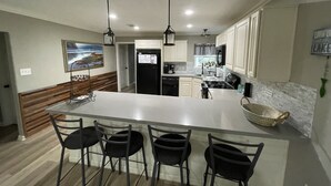 A modern kitchen with a large island, bar stools, pendant lighting, and a view of the living area. - !!!Private Lake!!! Fishing, Swimming & MORE!!! Charming Lakeside Bungalow!! (Conroe)