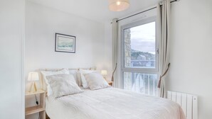 Et soveværelse med seng, natbord, lampe, et billede i ramme på væggen og et vindue med gardiner.