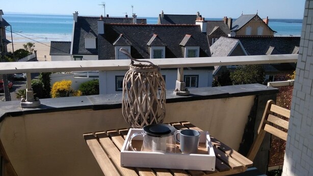 En balkon med et bord til to, med udsigt over en strand og boligbygninger.