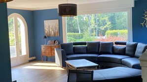 A modern living room with a sectional sofa, a small table, and a bookshelf. There is a large window with a view of trees and a garden.