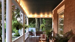 Un porche cubierto con suelo de madera, columnas blancas y un muro de ladrillo. Se observan dos sillas y una mesa con velas.