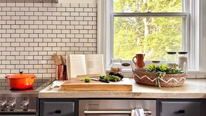 Una cocina con salpicadero de ladrillo, una ventana con vista a árboles, una estufa y diversos utensilios de cocina sobre la encimera.