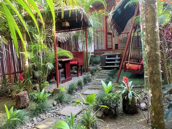 Un jardín tropical con un banco de madera, una hamaca y un sendero de piedra.