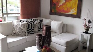 Een moderne woonkamer met een witte bank, een bijzettafeltje met decoratieve objecten, een schilderij aan de muur, en een raam met uitzicht op gebouwen.