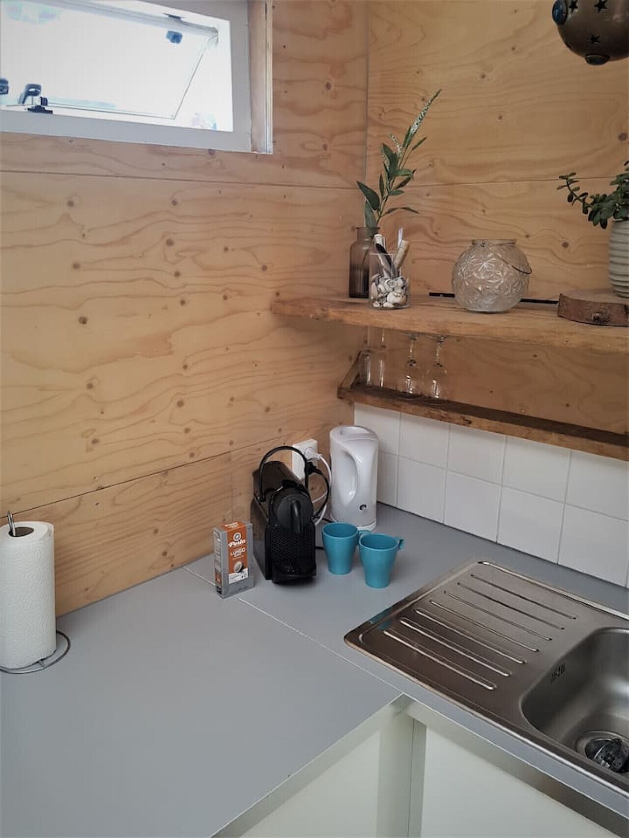 Une cuisine avec des murs en bois, un évier en acier inoxydable, une bouilloire, une machine à café et une étagère ornée d’objets décoratifs.