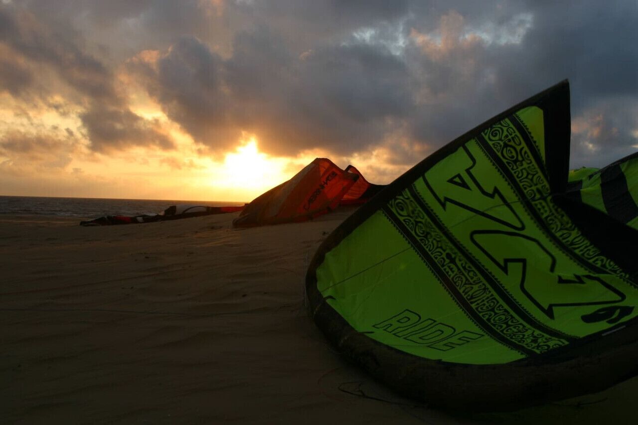Une plage avec une planche à voile posée sur le sable, une voile de kite-surf en arrière-plan et un coucher de soleil.