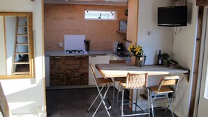 Cocina compacta con encimera de madera, una ventana pequeña, televisor y zona de comedor con sillas.